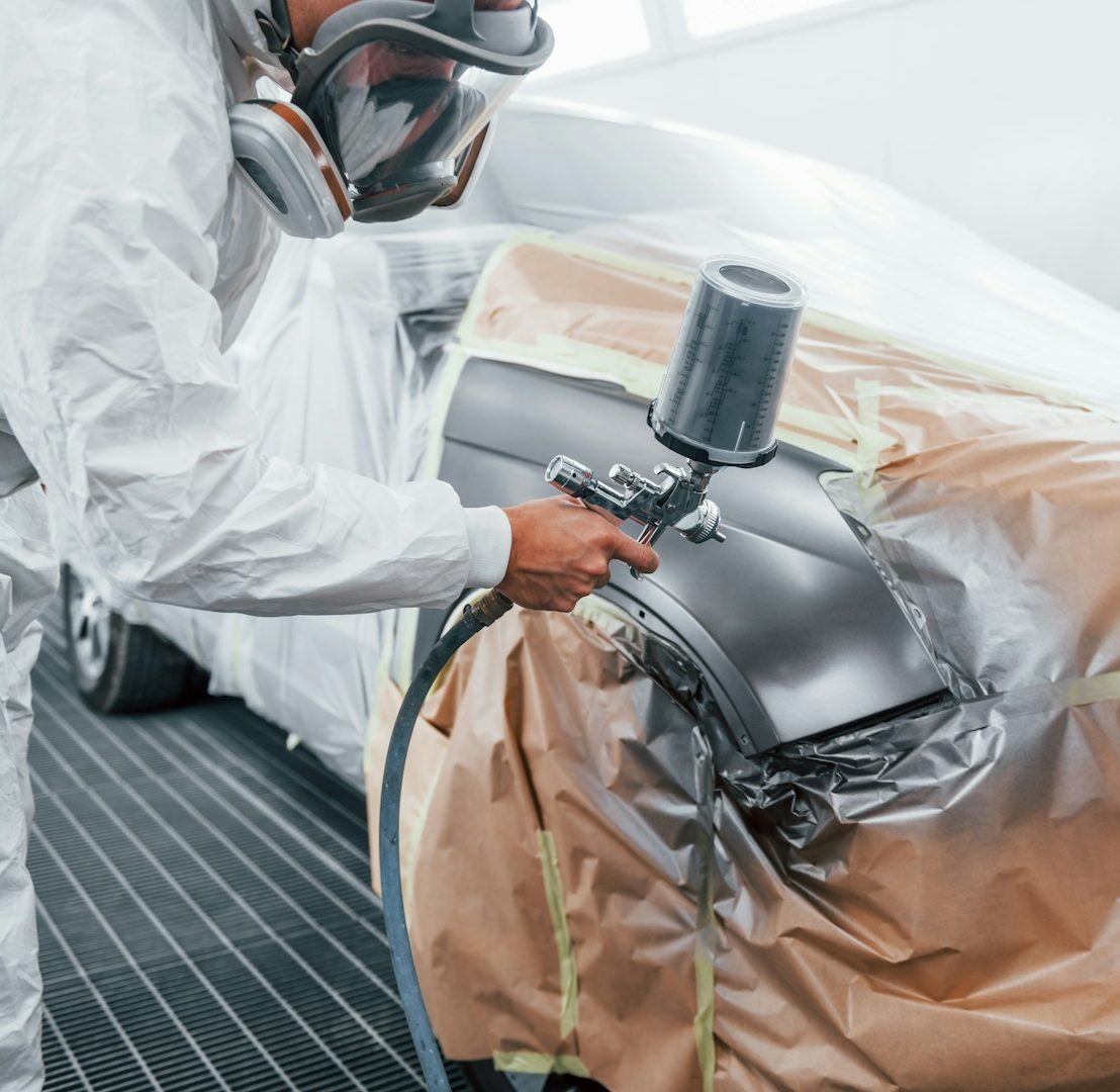 A person in protective gear spray-painting a car in a workshop.