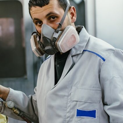 A person in a grey work coat and mask, preparing materials in a workshop setting.