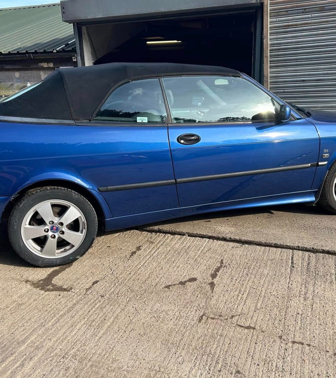 Blue convertible car parked at an industrial location with a garage in the background.